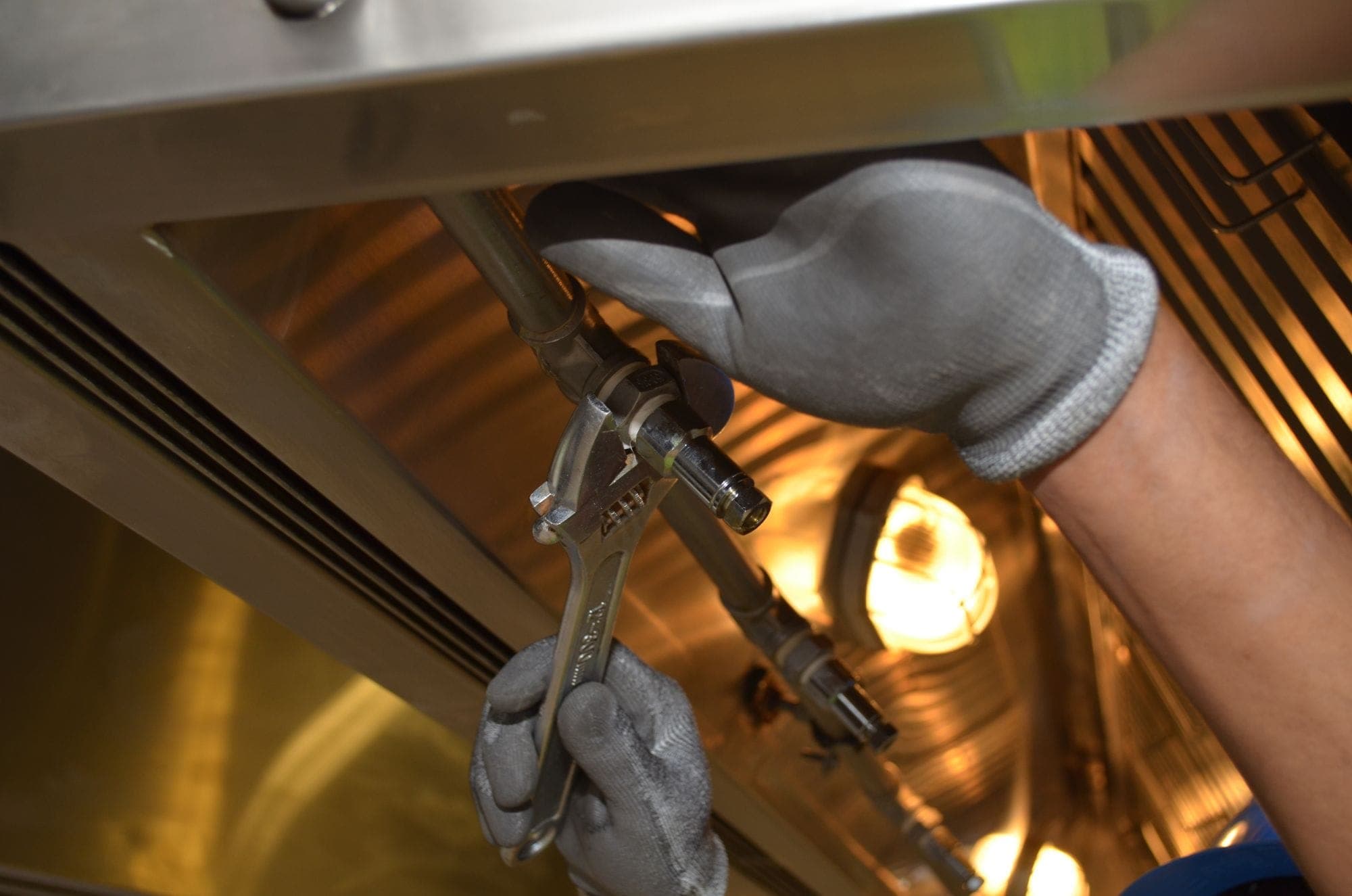 Gloved hands using an adjustable wrench on a gas pipe under a commercial kitchen hood